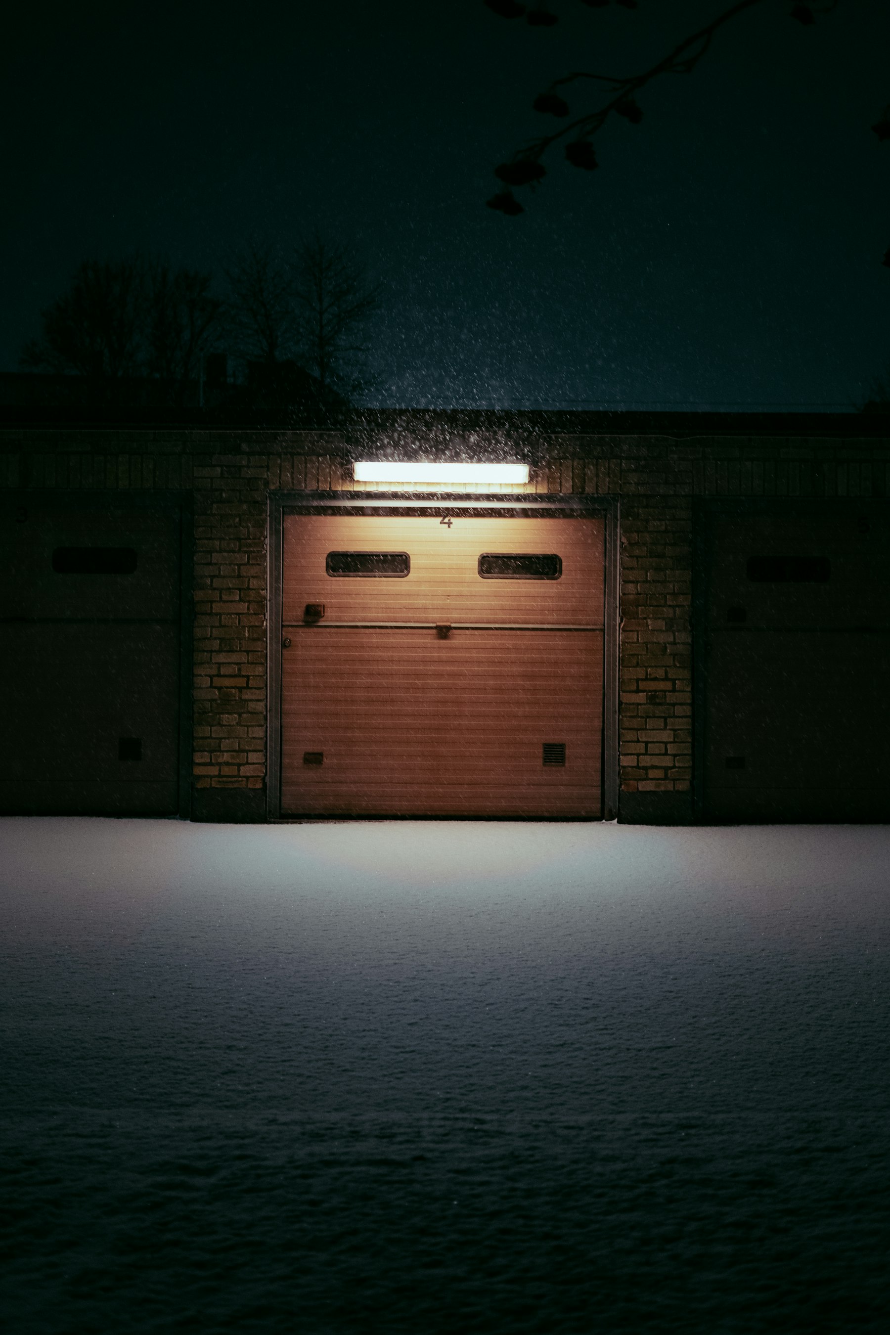 A roll up door lit by a single fixture at night, snow on the ground in front of it.