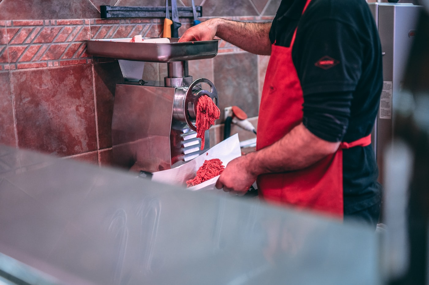 A butcher in an apron feeding trimmed meat through a stainless grinder onto paper.