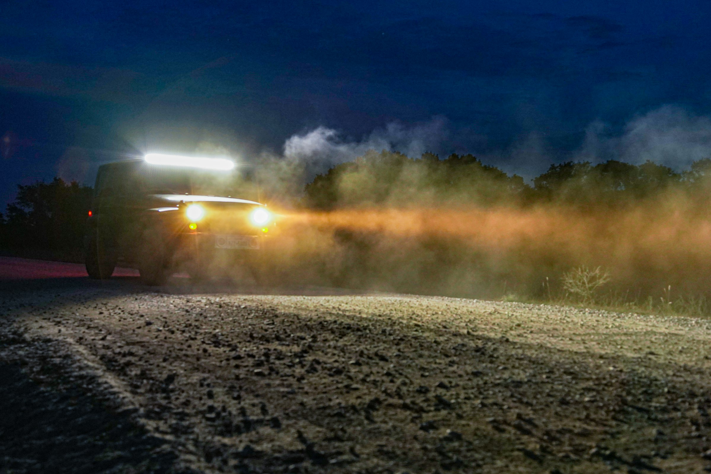 A truck on a gravel road after dark, headlights and a roof light bar throwing dust up into the beam.